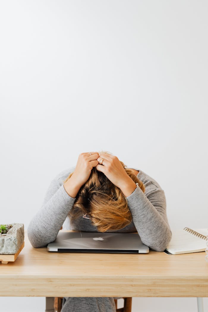 A woman in gray long sleeves resting her head on a laptop, appearing tired or stressed.